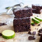 This image is used for decorative purposes. It features a stack of healthy chocolate brownies. There are chocolate chips in the foreground of the image and you can see slices of zucchini in the background.