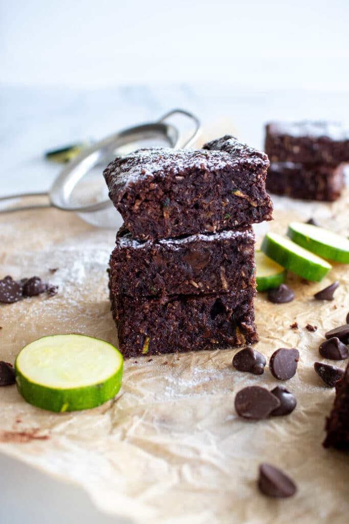 This image is used for decorative purposes. It features a stack of healthy chocolate brownies. There are chocolate chips in the foreground of the image and you can see slices of zucchini in the background.