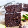 This image is used for decorative purposes. It features a stack of healthy chocolate brownies. There are chocolate chips in the foreground of the image and you can see slices of zucchini in the background.