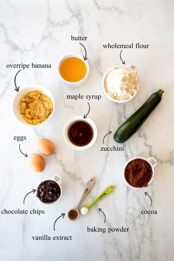 This image shows the ingredients needed for the chocolate zucchini brownies: mashed banana, butter, wholemeal flour, maple syrup, zucchini, cocoa, eggs, chocolate chips, vanilla extract, baking powder.